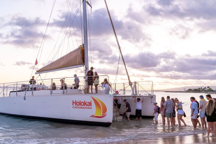 People boarding a catamaran named Holokai on a beach at sunset.
