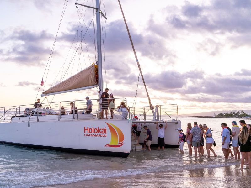 People boarding a catamaran named Holokai on a beach at sunset.