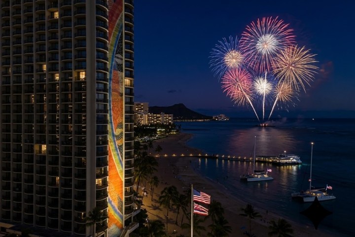 Fireworks over a city beach at night with lit building and ocean view. Holokai Catamaran